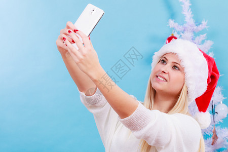 现代节日概念穿着圣诞礼帽的金发女人给自己拍圣诞照用智能手机自拍图片