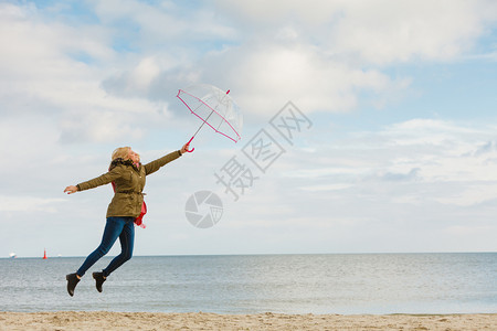 幸福享受寒冷的秋天气感受伟大的概念女人在海边滩上阳光明媚的白天和清蓝空上用透明的伞跳跃女人在海滩上用透明的伞跳跃图片