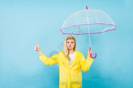 身穿黄色雨衣的金发女人拿着透明的雨伞检查天气是否雨下显示大拇指举手势图片