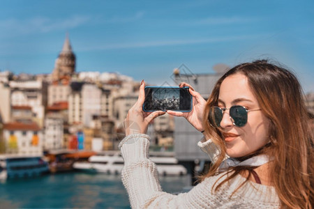 拥有耳机智能手和太阳眼镜的美丽迷人年轻女孩的肖像在土耳其伊斯坦布尔苏莱马尼耶清真寺Suleymaniye清真寺的加拉塔大桥上自拍图片