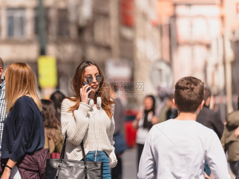 穿着带耳机和太阳眼镜的跳伞牛仔裤和的漂亮年轻女孩在人群中行走时使用智能电话图片