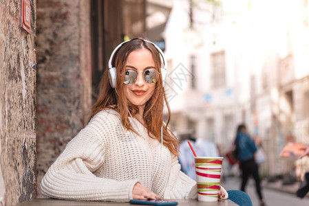 美丽的有魅力年轻时装女孩带着耳机智能手和太阳眼镜坐在街头咖啡厅喝果汁边坐着图片