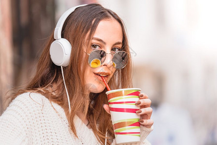 美丽的有魅力年轻时装女孩带着耳机和太阳眼镜在街头咖啡厅边喝果汁听音乐图片