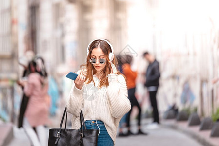 穿着跳伞和牛仔裤的美丽有魅力年轻时装女孩带着耳机智能手和太阳眼镜在人群中行走时聆听音乐图片