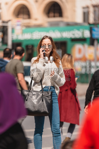 穿着带耳机和太阳眼镜的跳衣牛仔裤和的漂亮女孩在街上行走时用智能电话说图片