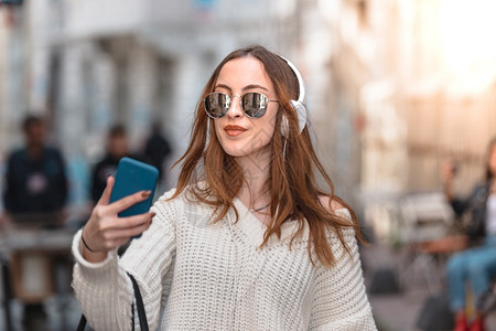 带着耳机智能手和太阳眼镜的跳跃式年轻时装女孩在人群中走动时会听音乐背景图片