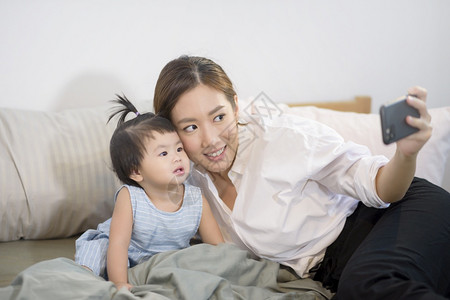 亚洲母亲及其女婴在床上家庭安全父母亲技术概念等场合向父亲打自拍或视频电话亚洲母亲及其女婴在床上技术概念等方面向父亲打自拍或视频电图片