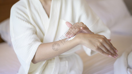 一位美丽的白浴袍女在卧室皮肤护理和治疗中施用体润湿乳霜图片