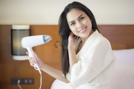 穿着白浴袍微笑的年轻女子在卧室洗完澡后用吹毛机擦发头干图片