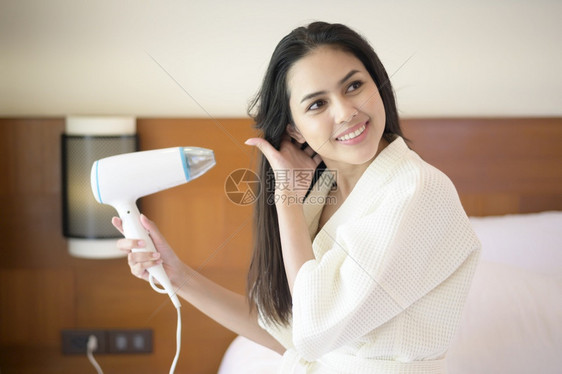 穿着白浴袍微笑的年轻女子在卧室洗完澡后用吹毛机擦发头干图片