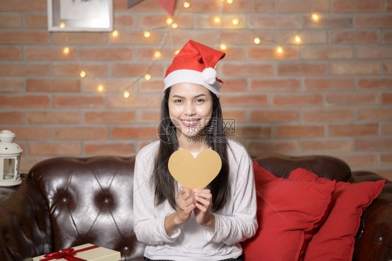 一个戴着红色圣诞老人帽子的微笑年轻妇女展示了圣诞节的心脏形状模型假日概念年轻微笑的女士穿着红色圣诞老人帽子节的心脏形状模型图片