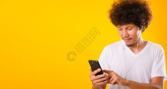 快乐的长着卷发亚洲帅男子他笑着享受听用黄色背景的移动智能手机隔离器在耳上听音乐图片