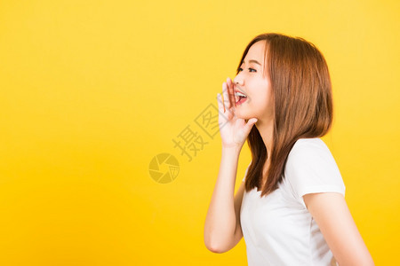 亚洲快乐肖像美丽的可爱年轻女孩站着穿T恤手在嘴上说悄的谣言试图与世隔绝摄影棚拍黄色背景复制空间图片