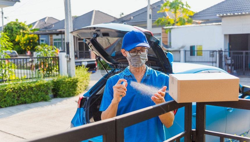 亚洲青年送货员网络上带一箱身穿制服的亚洲青年送货员他保护面罩和喷水洒净化根据宵禁检疫大流行冠状COVID19为家门口的顾客提供服图片