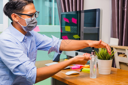在家庭办公室工作的亚洲商业青年男子他用防护面罩隔离疾病冠状毒或COVID19并在前台PC电脑上用清洁剂凝胶洗手图片