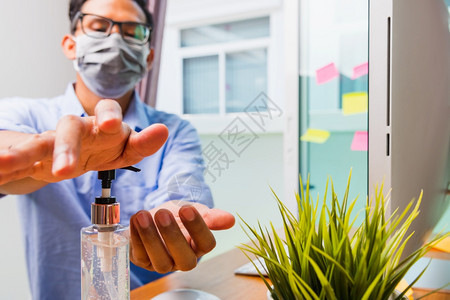 在家庭办公室工作的亚洲商业青年男子他用防护面罩隔离疾病冠状毒或COVID19并在前台PC电脑上用清洁剂凝胶洗手图片