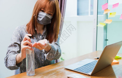 在家庭办公室工作的亚洲商业年轻女人他用防护面罩隔离疾病冠状毒或COVID19在前台笔记本电脑上用防污胶洗手图片