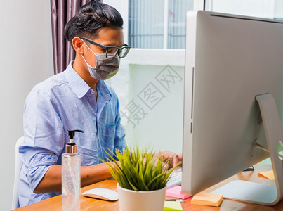 亚洲商人年轻男子身戴保护面罩和消毒凝胶在家庭办公室远程在线工作使用PC计算机检疫疾病冠状毒或COVID19图片