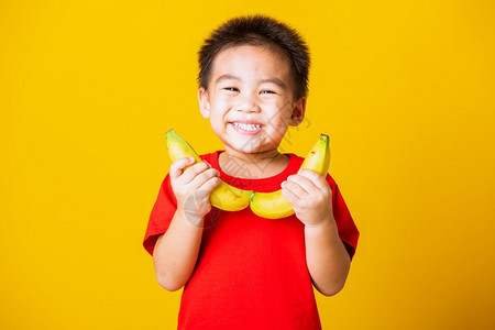 快乐的亚洲儿童或孩子可爱的小男孩带着红色T恤的笑脸穿着红色T恤玩香蕉水果摄影棚拍孤立的黄色背景图片