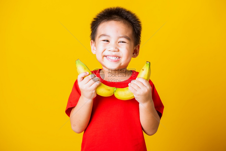 快乐的亚洲儿童或孩子可爱的小男孩带着红色T恤的笑脸穿着红色T恤玩香蕉水果摄影棚拍孤立的黄色背景图片