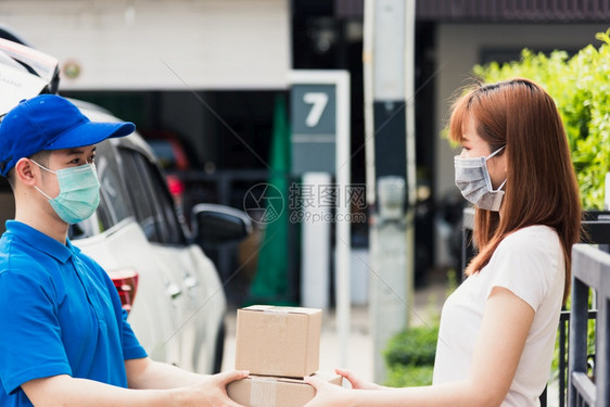亚洲青年送货员在线购买亚洲青年快递员提供包邮箱他保护面罩服务在宵禁检疫大流行冠状COVID19下在前家接受包箱的女客户图片