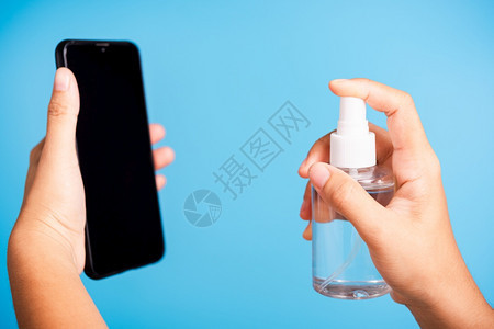 亚洲青年妇女将新闻撒布机净化剂酒精喷洒液射到智能移动电话清洗卫生预防COVID19或冠状保护概念孤立的蓝底背景图片
