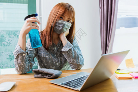亚洲商业女青年在家庭办公室工作他清洁笔记本电脑用清洁剂酒精喷洒隔离疾病冠状毒或COVID19在工作前佩戴保护面具图片