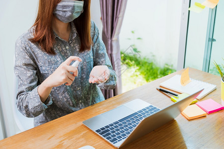 亚洲商业女青年在家庭办公室工作他用防护面罩隔离疾病冠状毒或COVID19在前台笔记本电脑上用洗净剂喷雾器手图片