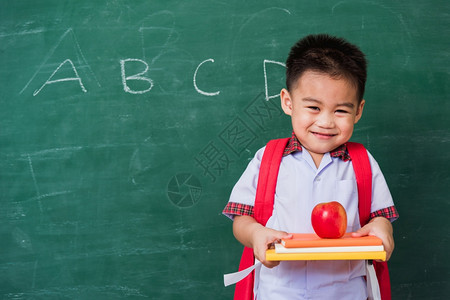 快乐的亚洲可爱小孩男从幼儿园的稚穿着学生制服拿书包红苹果在书上微笑绿色学校黑板上笑第一次到学校教育图片