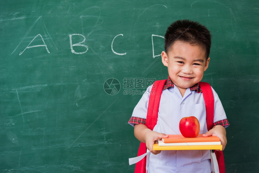 快乐的亚洲可爱小孩男从幼儿园的稚穿着学生制服拿书包红苹果在书上微笑绿色学校黑板上笑第一次到学校教育图片