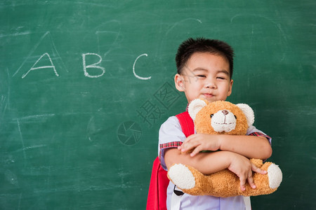 快乐的亚洲可爱小孩男从幼儿园穿着学生制服穿校带着书包微笑和拥抱泰迪熊在绿色学校黑板上第一次到学校教育图片