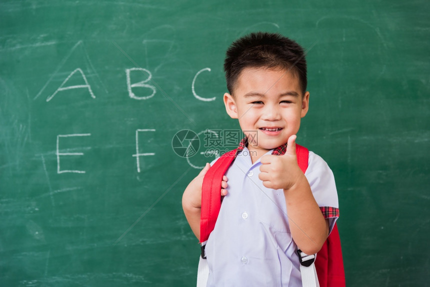 快乐的亚洲可爱小男孩幼儿园穿着校服带微笑的书包在绿色学校黑板上展示手指拇图片