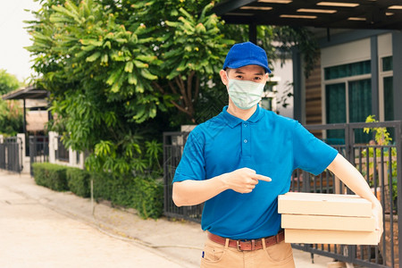 亚洲青年送货员运和持有快食披萨身穿制服面戴保护罩用手指着比萨盒根据宵禁检疫大流行冠状COVID19背景图片