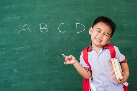 快乐的亚洲可爱小男孩幼儿园穿着学生制服书包和本在学校黑板上微笑指尖第一次上学图片