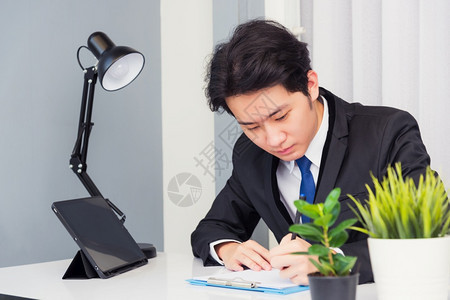 在家工作亚洲青年商人电视会议话或面对他笑着微地看用智能数字平板电脑坐在桌上的纸主办公室写笔记本资料图片
