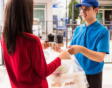 身穿蓝制服的亚洲年轻接生男青年他做杂货服务向根据流行冠状接受前门服务的妇女客户提供大米食品箱塑料袋回到新的正常概念图片
