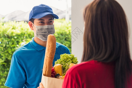 身穿制服的亚洲青年送货员戴保护面罩他制作杂货服务根据流行冠状向接受前门服务的女顾客提供新鲜食品回到的正常概念图片