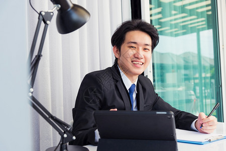 在家工作亚洲青年商人电视会议话或面对时他微笑地着看照相机用智能数字平板电脑坐在桌上家中办公室写笔记本资料图片