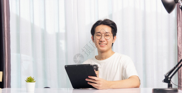 快乐的亚洲年轻商业英俊帅的在家办公室工作的人戴着眼镜T恤衫舒适他笑着用黑色现代智能数字平板电脑阅读子邮件或书桌上学习图片