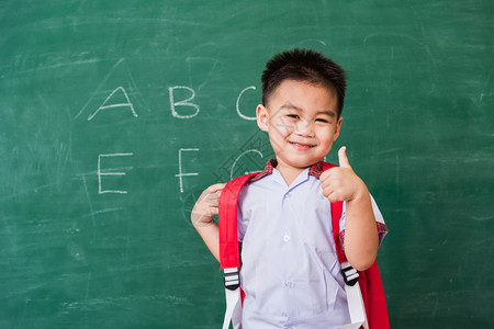 快乐的亚洲可爱小男孩幼儿园穿着校服带微笑的书包在绿色学校黑板上展示手指拇图片