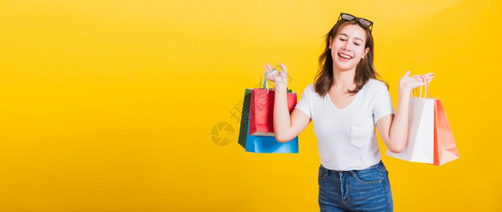 亚洲泰国人肖像快乐的美丽可爱年轻女子微笑着太阳镜兴奋地拿着购物袋满多色看着低头摄影棚拍着孤立的黄色背景带有复制空间图片