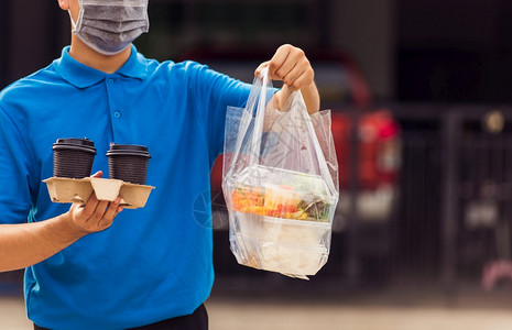 身着蓝制服的亚洲青年送货员戴面罩提供杂货服务根据流行冠状在前屋提供大米食品盒塑料袋和咖啡回到新的正常概念图片