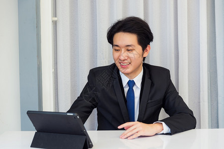 在家工作亚洲青年商人视频会议电话或面对时他微笑地着看照相机坐在桌子上用智能数字平板电脑触摸家庭办公室的屏幕图片