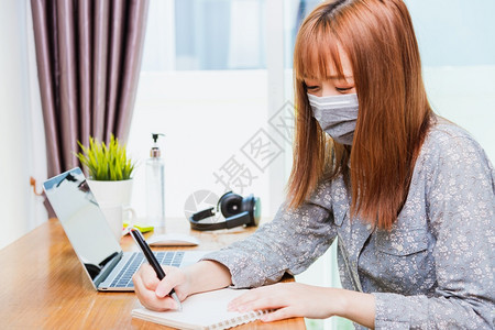 身戴面罩的亚洲年轻商业女人在家庭办公室用笔记本电脑工作用笔记本电脑隔离冠状或COVID19疾病并在今天写笔记簿工作图片