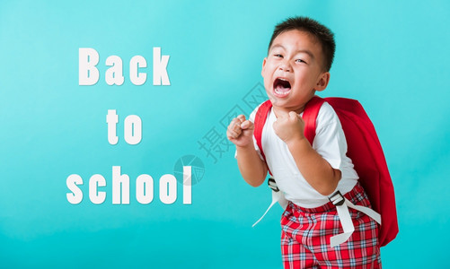 穿着制服笑脸的亚洲可爱男孩回到学校时举起手来欢庆孤立的蓝背景幼稚园时带着背包的孩子图片