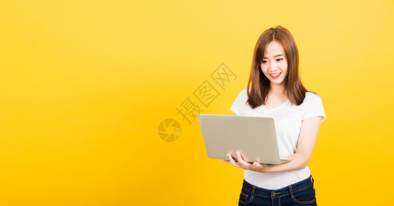 亚洲快乐肖像美丽可爱的年轻少女微笑着的站穿T恤衫使用笔记本电脑图片