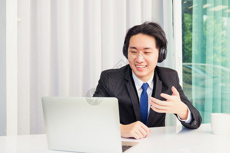 在家工作时亚洲青年商人微笑戴着耳机和西装微笑带着耳机和西装视频会议电话或面对在办公桌上并举手向在家办公的同事解释情况图片