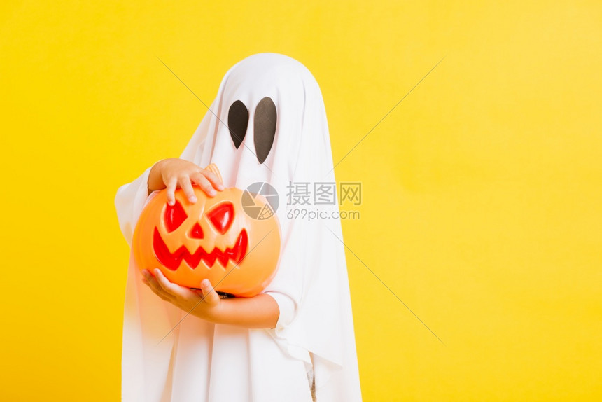 有趣的万圣节基概念可爱的小孩与白色服装扮神圣的灵吓他手握着橙南瓜的鬼摄影棚拍黄色的白背景高清图片下载 正版图片 摄图网