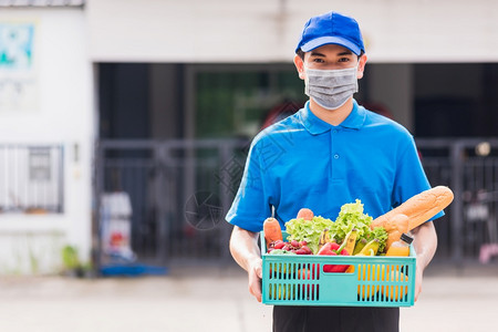 亚洲杂货店送员身穿蓝色制服面罩保护身穿蓝色制服在科罗纳爆发后在门前家的塑料箱中提供新鲜食品蔬菜回到新的正常概念图片