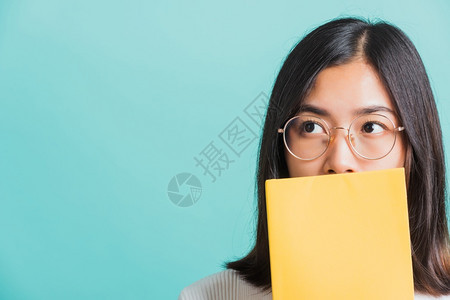 年轻美丽的亚洲女躲藏在一本公开的书背后身戴眼镜的肖像女拿着并阅读一本书蓝背景的摄影棚拍教育概念图片
